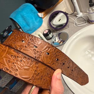 Può includere: Una cintura in pelle color cuoio con un motivo in rilievo di soli stilizzati, stelle e forme geometriche. La cintura presenta una finitura liscia e una fibbia metallica a un'estremità, con diversi fori circolari lungo la lunghezza.