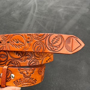 May include: A brown leather belt with embossed designs, including skulls and lightning bolts. The belt features the text "MARSHFIELD LEATHER CO." and a silver buckle. The belt is a medium brown color.