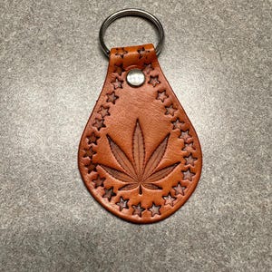 Op de afbeelding: Een bruine leren sleutelhanger met een zilveren sleutelring. De sleutelhanger heeft een gedetailleerd, gesneden marihuanabladontwerp, omringd door een cirkel van sterren.