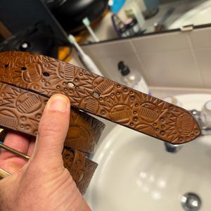 May include: A brown leather belt featuring a repeating embossed pattern of stylised suns and geometric shapes. The belt includes a series of oval-shaped adjustment holes and a metallic buckle, showcasing a detailed texture across the entire surface.