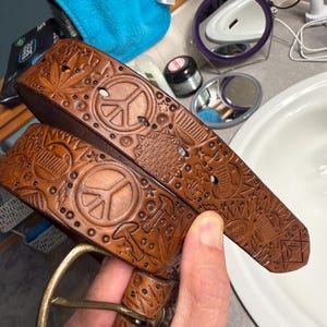 May include: A brown leather belt featuring intricate embossed designs including peace signs, mushrooms, cannabis leaves, and geometric patterns. The belt is equipped with a brass-toned metal buckle and has multiple punched holes for adjustment.