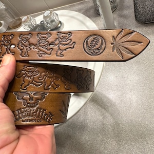 May include: Brown leather belt with a Grateful Dead inspired design. The belt features a skull, a marijuana leaf, and dancing bears. The text "Grateful Dead Leather Co." is embossed on the belt.