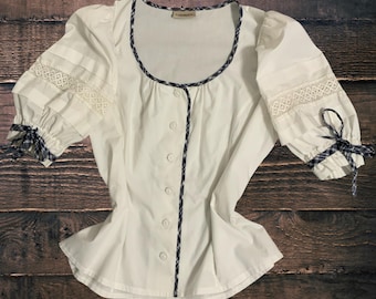 Pode incluir: Blusa branca com mangas curtas, decote redondo e fecho de botões. As mangas têm detalhes em renda e xadrez. A gola e a carcela dos botões são adornadas com um padrão xadrez azul escuro e branco.