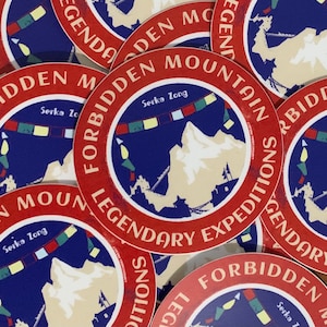 Puede incluir: Un conjunto de pegatinas redondas con borde rojo y la inscripción "FORBIDDEN MOUNTAIN LEGENDARY EXPEDITIONS" en blanco. El centro presenta un fondo azul con una cordillera y banderas de oración coloridas. También se incluye el texto "Serka Zong".