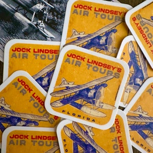 May include: Square coasters with a vintage design featuring a seaplane and the text "Jock Lindsey's Air Tours Florida" in red and blue on a yellow background. A black and white photo of a seaplane is in the background.