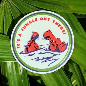 May include: A round sticker with a light green background and a green and blue border. The sticker features two red hippos in water, with the text "IT'S A JUNGLE OUT THERE!" in red. The sticker also includes the text "#ADVENTURERSPROJECT".