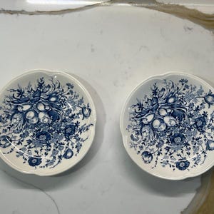 May include: Two white ceramic plates with a blue floral design. The plates have a scalloped edge and a detailed pattern of flowers and foliage. The plates are suitable for serving appetizers or desserts.