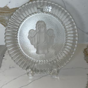 May include: Clear glass plate with a frosted angel design in the center. The plate has a scalloped edge and is displayed on a clear stand. A small sticker with the word "Goebel" is visible on the plate.