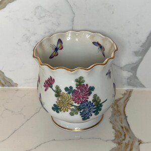 May include: White ceramic vase with a scalloped rim and gold trim. The vase is decorated with colorful floral and butterfly designs. The flowers are in shades of pink, yellow, and blue. The butterflies have purple, blue, and yellow wings.