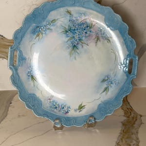 May include: A round, hand-painted serving platter with a light blue scalloped edge and handles. The white center features delicate blue floral designs with green leaves. The platter is supported by a clear stand.