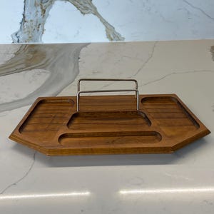 May include: A wooden serving tray with a geometric design and a stainless steel handle. The tray has several compartments and a warm, brown finish. The tray is designed for serving food or displaying items.