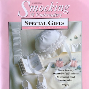 May include: A cream-coloured baby bonnet with lace trim and ribbon ties, alongside matching baby shoes and a small book. The image is from an Australian "Smocking & Embroidery" magazine, featuring gift ideas for smocking and embroidery.