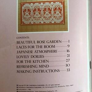 May include: A book page featuring a detailed illustration of white lace designs on a reddish-brown background. The image is framed within a gold border. The book's contents include topics like 'Beautiful Rose Garden' and 'Lovely Doilies'.