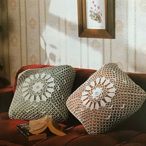 May include: Two square crochet pillows with floral center designs rest on a red sofa. The pillows feature a white floral pattern on a darker background, with an open, net-like crochet stitch. A book lies open nearby.