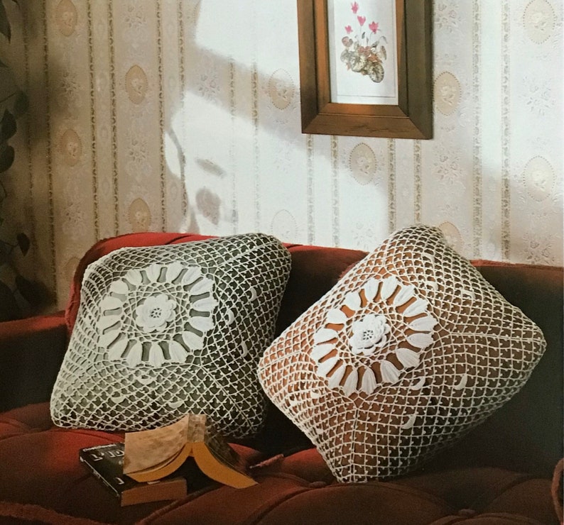 May include: Two square crochet pillows with floral center designs rest on a red sofa. The pillows feature a white floral pattern on a darker background, with an open, net-like crochet stitch. A book lies open nearby.