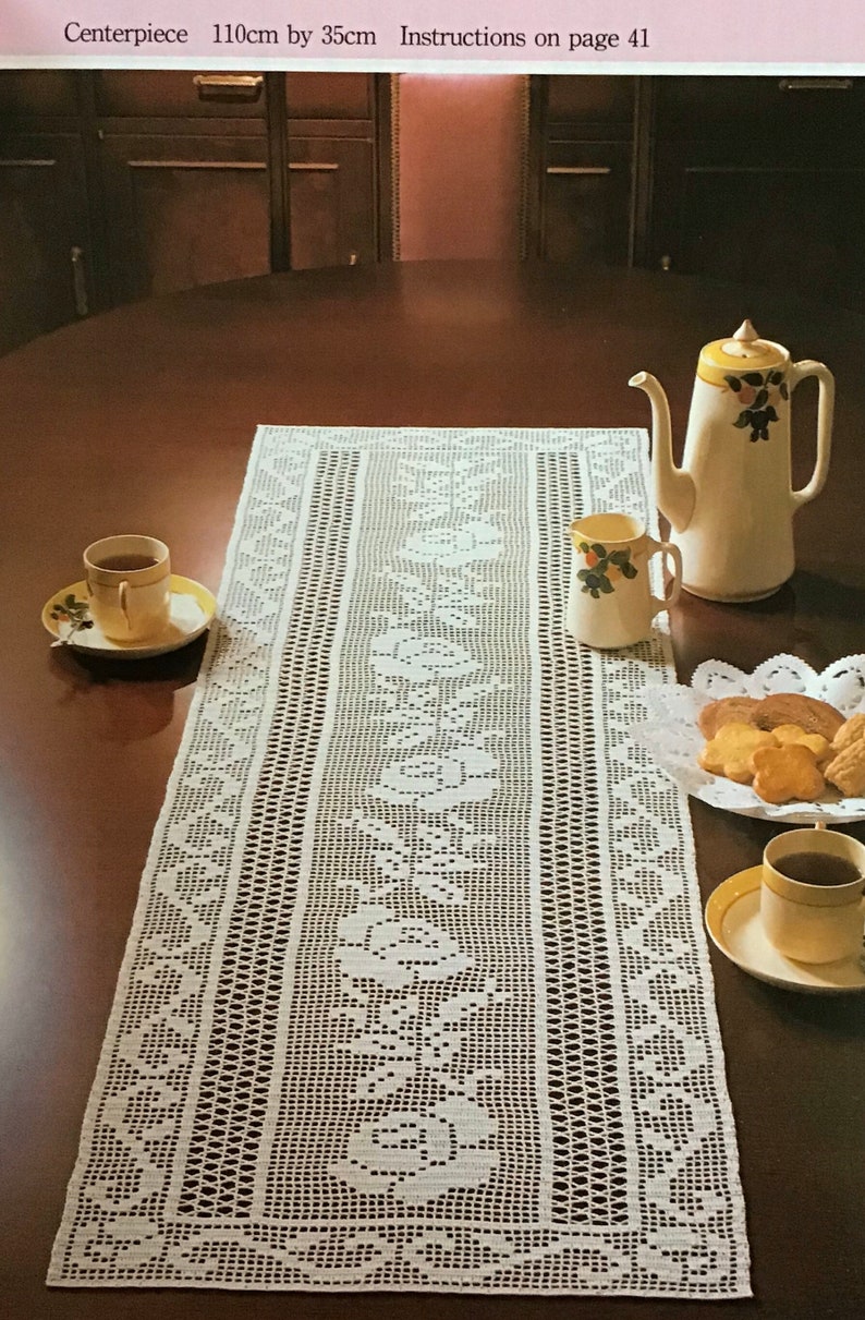May include: A white, open-weave crochet table runner, 43 inches long by 14 inches wide, with floral patterns. A teapot, creamer, teacups, and cookies are arranged on a dark wood table. The text "Centerpiece 110cm by 35cm" is visible.