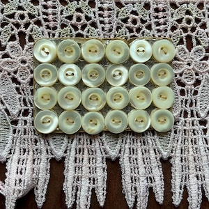 May include: A rectangular cardboard card holding twenty round, off-white, two-hole buttons arranged in a four-by-five grid. The buttons have a smooth, pearlescent finish and are positioned against a background of intricate, pale pink lace fabric.