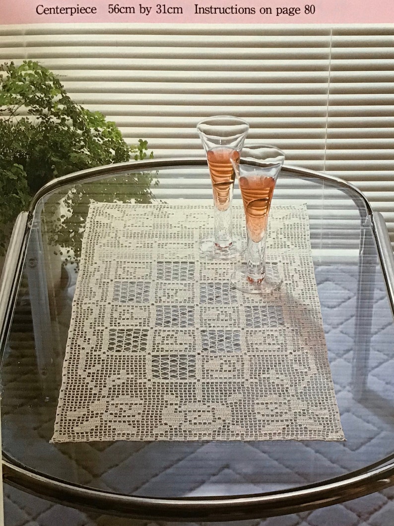 May include: A crocheted centerpiece, measuring 22 inches by 12 inches, is displayed on a glass table. Two stemmed glasses with a pink liquid sit atop the table. The text "Centerpiece" is visible.