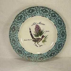 Può includere: Piatto decorativo con bordo turchese e dorato a fantasia. Il centro presenta un corridore, una pianta di yucca e le parole "New Mexico" e "Road-Runner". Il piatto ha un centro color crema.
