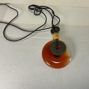 Könnte beinhalten: Eine Anhängerkette mit einem großen, runden, orange-weißen Stein, einer dunklen Münze und einem kleinen geschnitzten Knochenakzent. Die Kette hat eine schwarze Kordel.