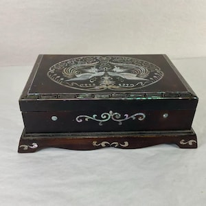 May include: A dark brown wooden box with a decorative lid featuring a mother-of-pearl inlay design of two birds. The box has a matching base and additional mother-of-pearl accents on the front. The box is rectangular and appears to be a jewelry box.