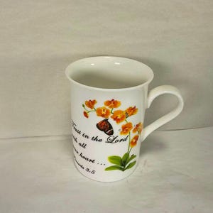 May include: White ceramic mug featuring a floral design with orange flowers and a butterfly. The mug has the text "Trust in the Lord with all your heart... Proverbs 3:5". The mug has a curved handle.