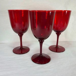 May include: Three ruby red wine glasses. Each glass has a rounded bowl, a slender stem, and a circular base. The glasses are made of a transparent material, allowing light to pass through. The glasses are arranged on a white surface.