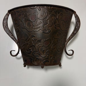 May include: A dark brown metal wall planter featuring an embossed floral pattern across its surface. The vessel has a wide, open top with a rope-textured rim, two curved side handles, and a flat base supported by small, rounded metal feet.