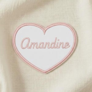Könnte beinhalten: Ein herzförmiger Aufnäher mit weißer Mitte und rosa Rand. Das Wort "Amandine" ist in rosa Schrift über die Mitte gestickt. Der Aufnäher befindet sich auf einem cremefarbenen, strukturierten Stoff.