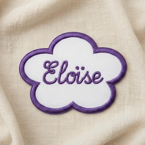 Könnte beinhalten: Ein weißer, wolkenförmiger Aufnäher mit lila Rand und dem Namen "Eloïse" in lila Schrift gestickt. Der Aufnäher liegt auf einem cremefarbenen Stoffhintergrund.
