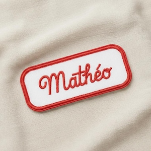 Könnte beinhalten: Ein weißes Namensschild mit rotem Rand und dem Namen "Mathéo" in roter Schrift gestickt. Das Schild ist rechteckig mit abgerundeten Ecken und vor beigem Stoffhintergrund.