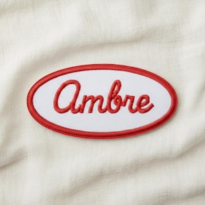 Könnte beinhalten: Ovaler Aufnäher mit dem Namen "Ambre" in roter Schrift auf weißem Grund, umrandet von einem roten Rand. Der Aufnäher befindet sich auf einem cremefarbenen Stoff.