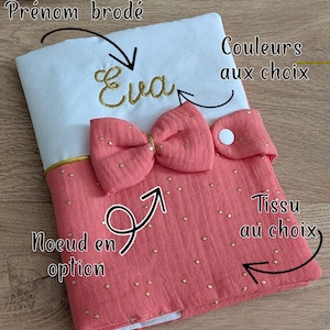 May include: Embroidered baby book cover with the name "Eva" in gold thread. The cover has a pink bow and measures approximately 22 cm x 17 cm. The design includes options for fabric and colours. The cover is padded.