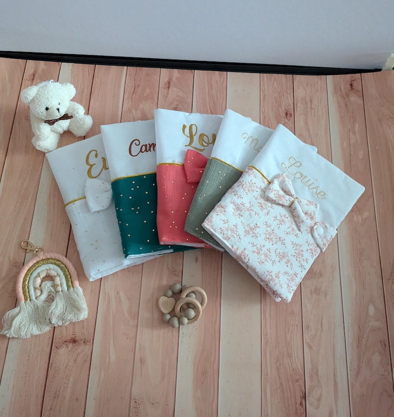 May include: Five personalised baby blankets with names embroidered in gold. The blankets feature various colours and patterns, including floral and polka dots. A white teddy bear, a rainbow keychain, and a wooden teether are also visible.