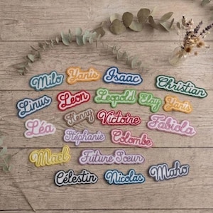 Peut inclure: Une collection d'écussons brodés avec des prénoms dans diverses couleurs et polices cursives, incluant Milo, Yanis, Isaac, Christian, Linus, Leon, Leopold, Elay, Henry, Victoire, Fabiola, Léa, Stéphanie, Colombe, Mael, Future Sœur, Célestin, Nicolas et Maho.