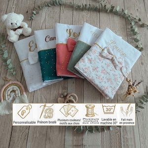 Peut inclure: Cinq housses en tissu rectangulaires avec prénoms brodés, nœuds décoratifs et motifs variés incluant des fleurs, des pois et des unis. Le texte indique Personnalisable, Prénom brodé, Plusieurs couleurs/motifs aux choix, Lavable en machine 30°, Fait main en provence.