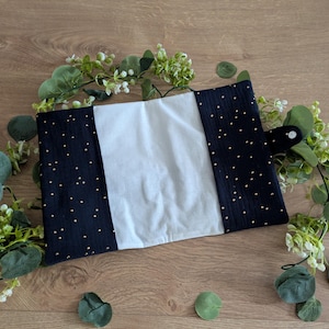 May include: A handmade book cover featuring a central white panel, framed by navy blue fabric dotted with gold specks. It has a black button closure. The cover is surrounded by green foliage, giving a natural look.