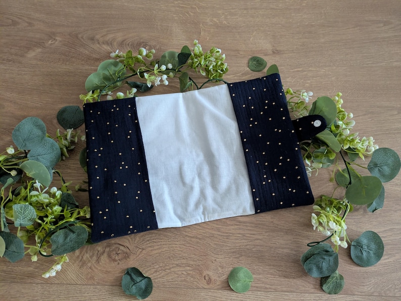 May include: A handmade book cover featuring a central white panel, framed by navy blue fabric dotted with gold specks. It has a black button closure. The cover is surrounded by green foliage, giving a natural look.