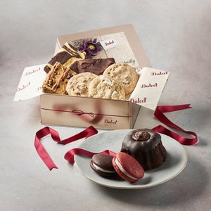 May include: A gift box filled with assorted cookies and brownies, including chocolate chip, peanut butter, and chocolate fudge. The box is tied with a red ribbon and the lid has the words "Dulcet Gift Bakery" printed on it. The box is sitting on a gray surface with a red ribbon and a plate of cookies and brownies.