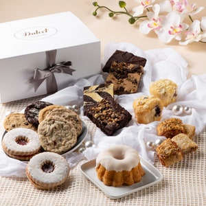 May include: A white gift box with a gray ribbon and a label that says "Dulcet" is surrounded by a variety of baked goods, including cookies, brownies, muffins, and a cake.
