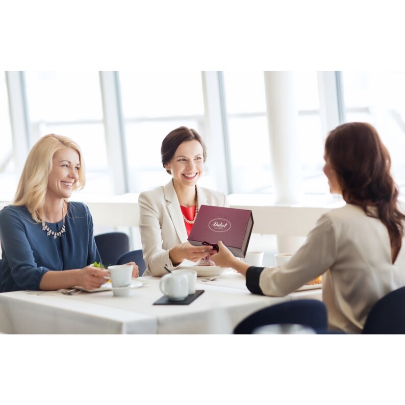 May include: Three women are sitting at a table, one is handing a burgundy gift box with a white ribbon and the text "Dukat" to another woman.