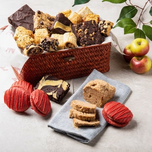 May include: A wicker basket filled with a variety of baked goods, including brownies, cookies, and pastries. The basket is sitting on a table with a gray tablecloth and red and green apples.