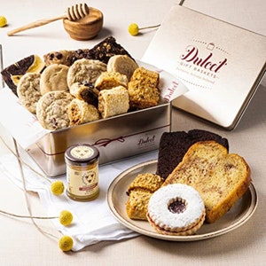 May include: A silver tin filled with a variety of cookies and pastries, including chocolate chip cookies, shortbread cookies, and biscotti. The tin is labeled "Dulcet Gift Baskets" and has a red ribbon tied around it. A small jar of honey is also included in the image.