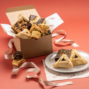 May include: A brown cardboard box filled with a variety of cookies and pastries, including hamantaschen, rugelach, and chocolate chip cookies. The box is sitting on a pink surface with a white plate containing two hamantaschen cookies.
