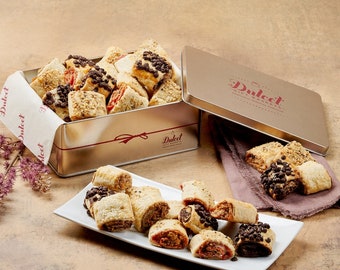 Puede incluir: Una lata de plata llena de galletas enrolladas surtidas, con una etiqueta roja y blanca que dice "Dulcel Gift Baskets". Las galletas están dispuestas en un plato blanco, con algunas que se caen de la lata.