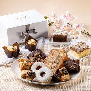 May include: A white gift box with a silver ribbon sits next to a white platter filled with various baked goods. The platter includes muffins, cookies, brownies, and slices of cake. The word "Dulcet" is printed on the box.