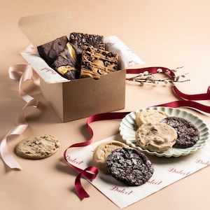 May include: A brown cardboard box filled with assorted brownies and a plate of chocolate chip, oatmeal raisin, and chocolate cookies. The box is tied with a pink ribbon and the cookies are on a white plate with a scalloped edge.