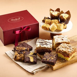 May include: A red gift box with a ribbon bow, and an assortment of baked goods. Brownies, muffins, and bars are arranged on a wooden board, a plate, and a linen napkin. The box has the text "Dulcet Bakery" in gold.