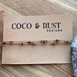 May include: A thin brown cord bracelet featuring small, multicoloured beads spaced along the length. The bracelet is displayed on a rectangular tan card printed with the black text COCO & RUST DESIGNS, positioned next to a large, jagged purple amethyst crystal.