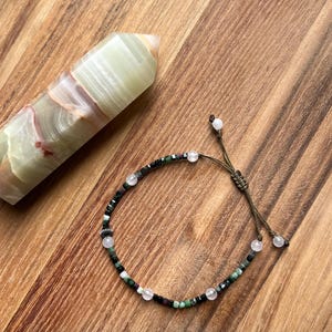 May include: A beaded bracelet featuring small green, black, and red stone beads interspersed with light pink round beads and a single silver-toned metal spacer bead, finished with a brown cord and an adjustable sliding knot closure mechanism.